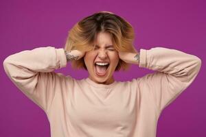 Indoor portrait of young dyed blonde female, with tattooed arms keeps her hands on head, screaming with angry facial expression, isolated over purple background photo