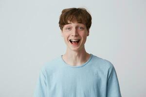 Close up of amazed excited young man with short disheveled hair and braces on teeth wears blue t-shirt shouting and feels happy surprised isolated over white background photo