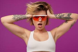 Indoor portrait of young dyed blonde female, with tattooed arms wears bright orange glasses, keeps her mouth opened with shocked facial. expression. isolated over purple background photo