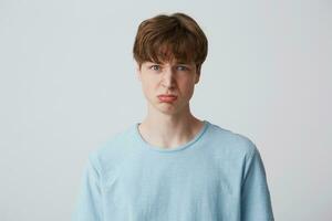 Portrait of blue-eyed young guy looks sad, upset, frustrated, displeased with the result of the test, lips pout, expresses insult, wears blue t-shirt stands isolated over white background. photo