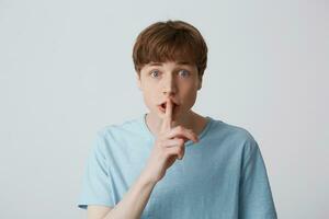 Young guy with wide open eyes, keeps fore finger on lips, calls to keep a secret, do not tell anyone, keep quiet, do not make noise, demonstrates silence gesture, isolated over white background photo