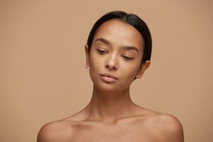 hermosa caucásico chica, mirando abajo, después un protección de la piel y spa, con sano limpiar Fresco piel. bonito mujer con negro pelo fijo detrás, grueso Cejas y desnudo espalda terminado beige antecedentes foto