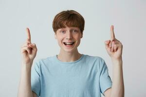 Boy is fascinated and excited. Portrait of amazed attractive young guy in blue t-shirt pointing with index fingers up and looking camera with wide smile, showing braces on teeth, over white wall photo