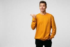 Indoor photo of happy glad handsome guy,looks amazed excited, popping eyes out dressed in orange sweatshirt, points with thumb finger on a blank white wall with copy space for your promotional content