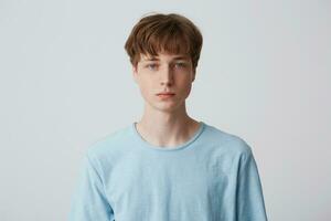 Close up face of a young man without emotions. Beautiful emotionless guy in a blue t-shirt looking to the camera, isolated over white background photo