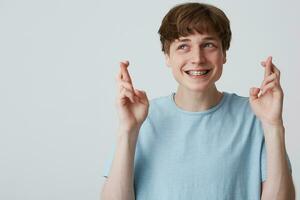 Portrait of young guy looks away making a wish, hoping for something pleasant, hands up and fingers twisted, with braces on teeth, dressed casually,blank copy space for your promotional content photo