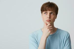 Thoughtful focused european young man in blue t-shirt, keeps arm on chin and looking distance, furrowed eyebrows, being confused and questioned, thinking what do to have fun, troubled, over white wall photo