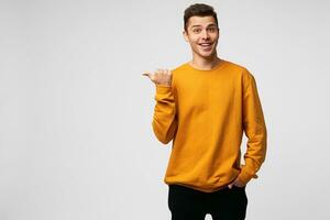 Young attractive handsome guy looks delighted gladden amazed, mouth open, smiling, like dream came true, big discount in shop,wears orange sweatshirt, points with thumb finger on a blank copy space photo