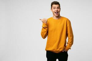 Young attractive handsome guy feels delighted gladden amazed,mouth open,smiling,wears orange sweatshirt, points aside on something incredible with thumb, blank copy space for promotional content photo