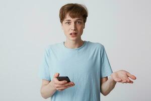 Guy with braces on teeth is looking straight in disbelief, holding a phone in his hand, the second hand is stretched forward. Mouth slightly open from indignation, feels injustice, shocked by passing photo