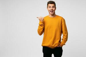 Indoor photo of funny happy delighted handsome guy, smiles, shining from happiness, completely satisfied, dressed in orange sweatshirt, points with thumb finger on a blank copy space