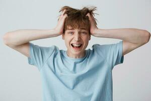 Screaming amazed excited young man clutched at his head, hair tousled, mouth wide opened as shouting loud with braces on teeth wears blue t-shirt, feels happy surprised isolated over white background photo