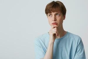 Focused young man in blue t-shirt, keeps arm on chin and looks thoughtfully away, being confused and questioned, thinking what do to solve the problem, troubled, over white background photo