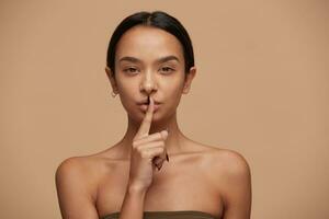 Young mixed race girl calls for silence, do not tell anyone,keep quiet, do not make noise, keeps fore finger on lips,demonstrates silence gesture,head and shoulders,on beige background photo