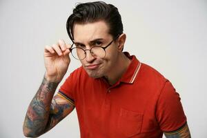 Headshot of angry male looks seriously at camera, frowns face, touches rim of glasses, looks in bewilderment, isolated over white background. Photo of annoyed man stares through spectacles