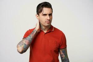 Macho, keeps his hand on his nape, looks askance, his eyebrows frowned. Waist up shot of black hair young man, with styling,in a red polo t-shirt, has tattoos, on a white background photo