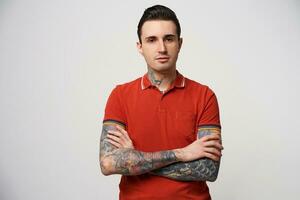 Young brunet man with his hair folded and normal confident facial expression, wearing a red polo t-shirt stands with his arms crossed, has a tattoo on his arms and neck, over white background photo