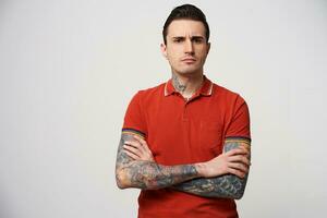 Serious man with arms folded look suspicious, eyebrows frowning, has a tattoo on his arms and neck, standing isolated on a white background and looking at camera photo