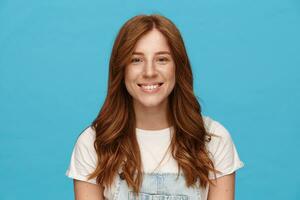 Portrait of young pretty happy lady with foxy wavy hair looking cheerfully at camera with wide smile and keeping hands down while posing over blue background photo