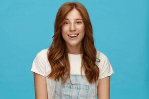 Joyful young charming redhead woman with natural makeup looking gladly at camera with broad sincere smile, being in nice mood while posing over blue background photo