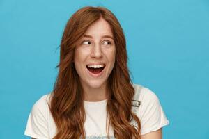 Joyous young beautiful redhead woman with wavy hairstyle looking cheerfully aside with wide smile, wearing basic white t-shirt while standing over blue background photo