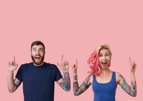 Overjoyed young attractive couple in casual blue shirts showing joyfully upwards with raised forefingers, rounding their eyes and keeping mouthes wide opened, isolated over pink background photo