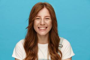 Close-up of young attractive green-eyed redhead woman showing her pleasant emotions and looking at camera with charming smile, isolated over blue background photo