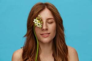 de cerca de agradable mirando joven positivo pelirrojo dama con blanco flores en su cara sonriente suavemente mientras en pie terminado azul antecedentes con cerrado ojos foto