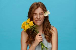 Portrait of young flirty lady with wavy hairstyle holding bouquet of flowers and biting underlip while looking playfully at camera, isolated over blue background photo