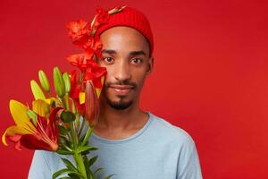 cerca arriba de joven frio hombre en rojo sombrero y azul camiseta, sostiene un ramo de flores en su manos, mira a el cámara con calmante expresión, soportes terminado rojo fondo foto