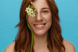 Romantic young lovely redhead woman with curls showing her pleasant emotions and smiling pleasantly at camera, standing over blue background with naked shoulders photo