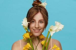 Indoor photo of glad young redhead female with bun hairstyle looking at camera with pleasant smile while standing over blue background with bouquet of flowers