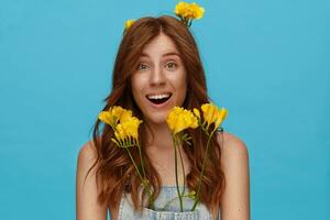 Exited young lovely lady with natural makeup raising surprisedly eyebrows while looking joyfully at camera, standing over blue background with yellow freesia photo