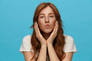 Portrait of young pretty redhead woman with curls folding her lips in air kiss and keeping hands raised while looking at camera, isolated over blue background photo