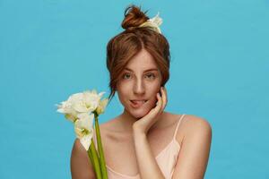 Indoor photo of young redhead green-eyed lady holding raised hand on her face and biting underlip while looking gently at camera, standing over blue background