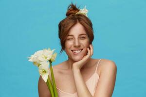 Positive young redhead attractive lady with bun hairstyle biting underlip while looking gladly at camera, holding bunch of white flowers while posing over blue background photo