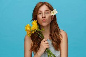 Indoor shot of young lovely green-eyed redhead woman with yellow flowers folding lips while looking positively at camera, standing over blue background in jeans top photo