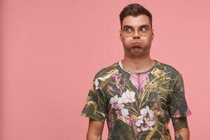 Headshot of young man grimacing, inflating his cheeks, holding his breath, looking aside with funny face expression, standing over pink background photo