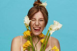 Funny shot of young attractive redhead lady with bun hairstyle showing cheerfully her tongue while fooling, standing over blue background with spring flowers photo