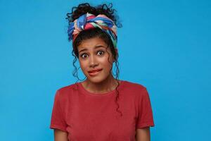 Portrait of confused young pretty curly female with gathered hair biting worringly underlip while looking perplexedly at camera, standing over blue background photo