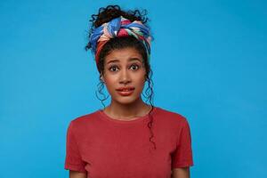 Bewildered young attractive dark haired curly woman with multi-colored headband frowning eyebrows while looking confusedly at camera, isolated over blue background photo