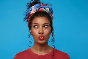 Close-up of young attractive dark haired lady with curls dressed in colored clothes looking aside while pouting her lips, standing over blue background photo