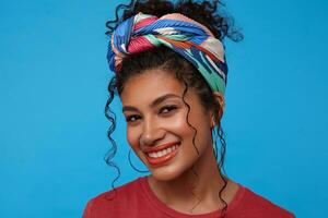 Close-up of young pretty dark haired lady with orange lips wearing multi-colored headband while posing over blue background, looking gladly at camera with broad smile photo