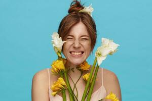 Studio shot of young joyful redhead female with bun hairstyle frowning happily her face while smiling widely, isolated over blue background with bunch of flowers photo