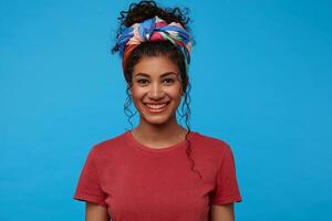 Portrait of young pretty brunette curly woman with festive makeup looking cheerfully at camera with charming smile, standing against blue background photo