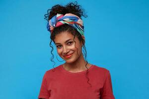 Glad young pretty dark haired curly lady with festive makeup dressed in burgundy t-shirt smiling gently while looking pleasantly at camera, isolated over blue background photo