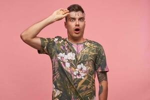 Indoor shot of young man in flowered t-shirt with glasses on his forehead, standing over pink background, looking to camera shocked and dazed photo