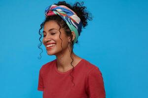 Good looking happy young brunette curly woman with multi-colored headband being in nice mood and miling gladly with closed eyes while standing over blue background photo