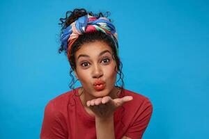 Indoor shot of positive young brown-eyed brunette curly female keeping palm raised while blowing air kiss at camera, standing over blue background in colored clothes photo