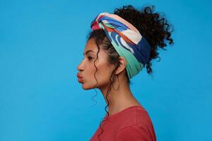 Studio photo of young dark haired curly woman with multi-colored headband folding her lips in air kiss while looking ahead, standing over blue background